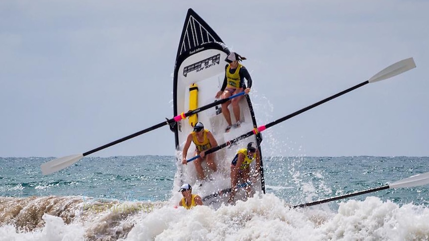 A surfboat leaps vertically out of rough surf with four women at the oars