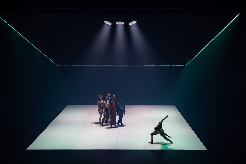 Six dancers stand in formation on a white-floored stage, while another dancer in the corner lunges with his arms behind him