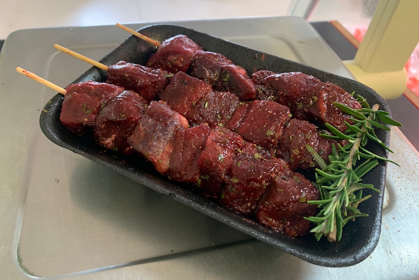 A black tray with three emu meat kebabs on skewers and a sprig of rosemary.  
