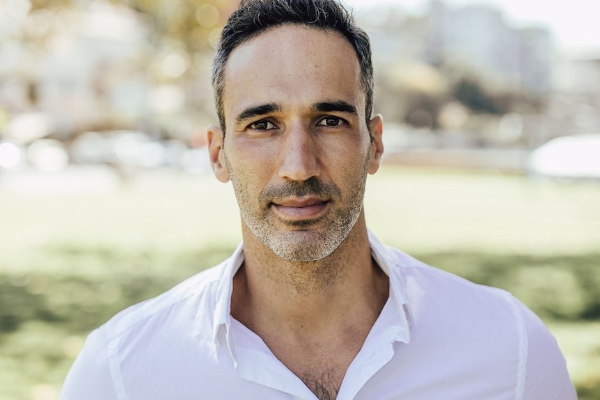 A photo of Alex wearing a white shirt with trees in the background.