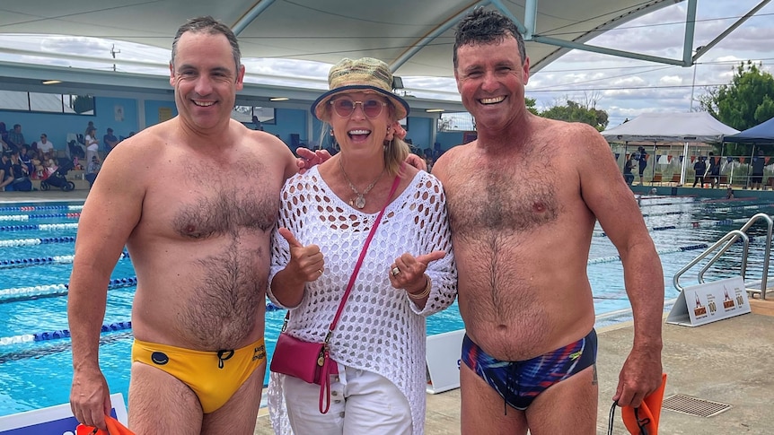 Two men wearing swim wear and a woman stand in a line smiling in front of a lane pool