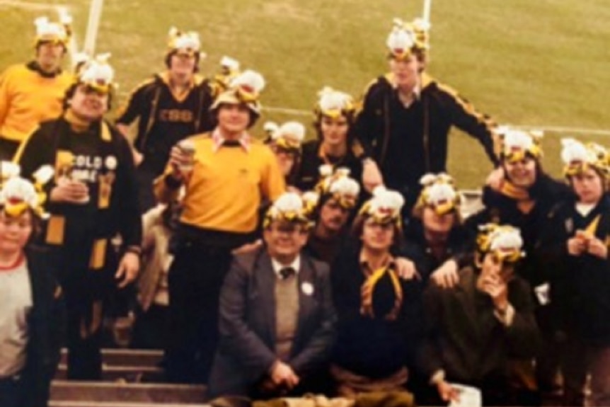 A grainy, old photograph shows a group of people gathered in the stands of a football oval dressed in yellow and black.