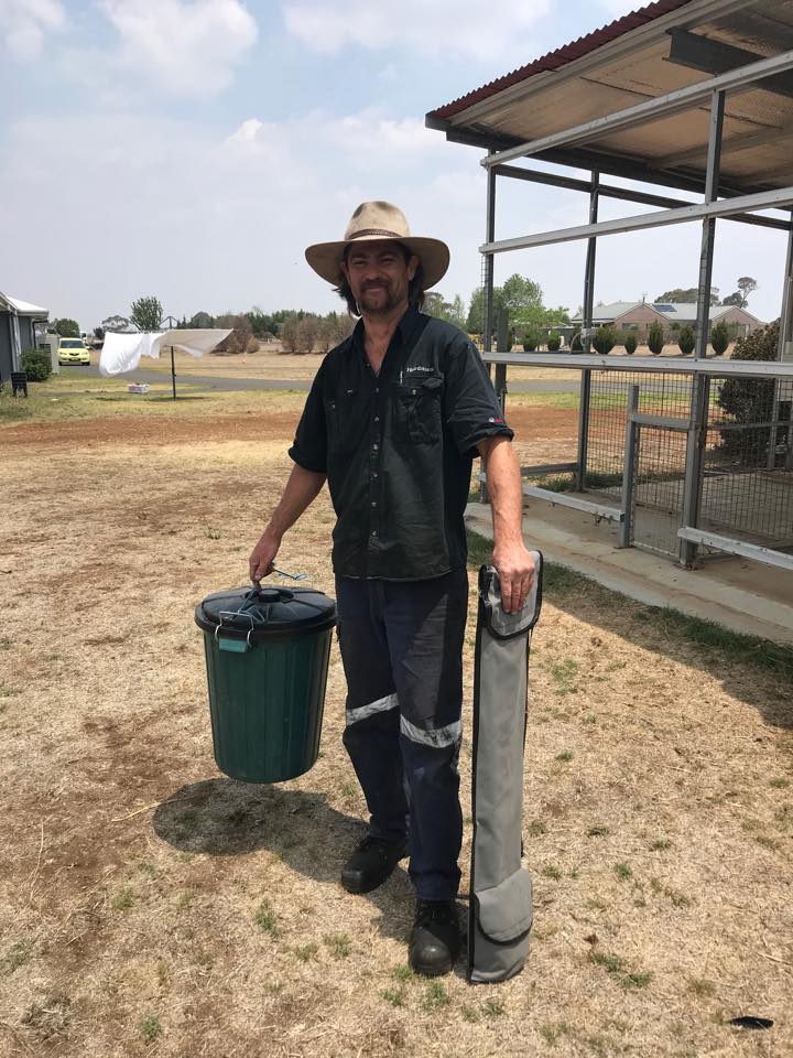 A man wearing all black and a hat, holds a bin and snake catch whilst smiling at the camera.