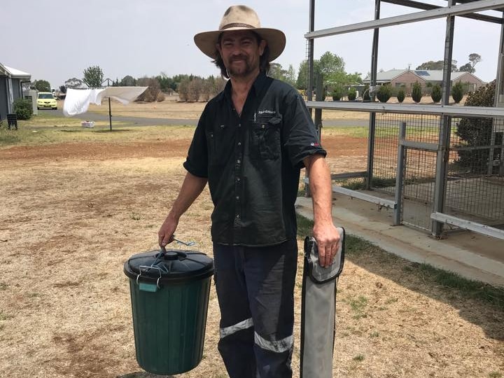 A man wearing all black and a hat, holds a bin and snake catch whilst smiling at the camera. 