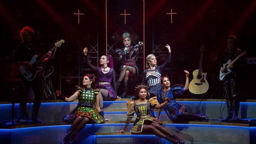 Six women in costume sit in the spotlight on stage with band members