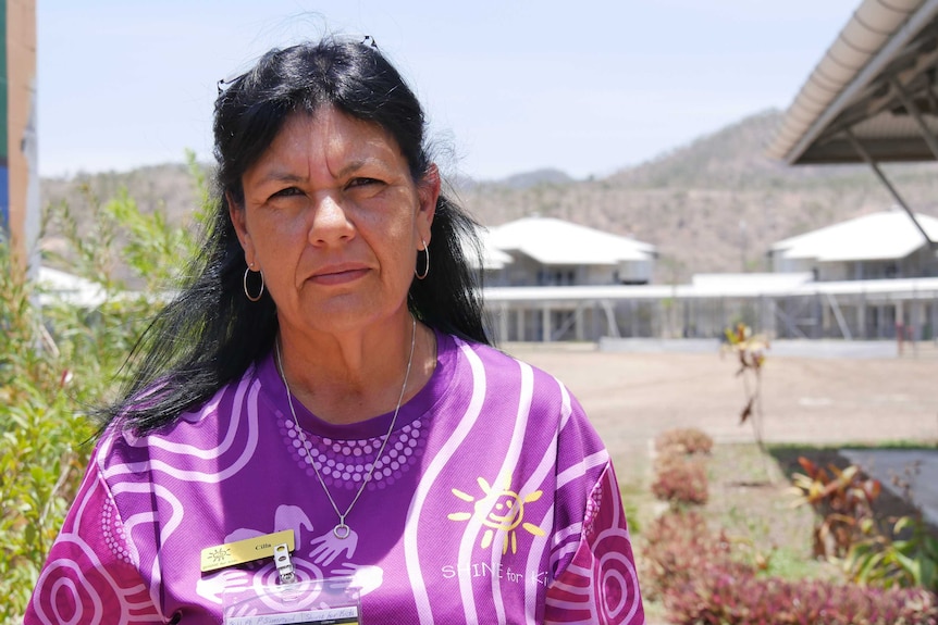 Woman looks at camera, Indigenous, long hair, wearing bright purple TShirt and hoop earrings, inside prison