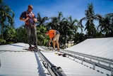 Two men standing on a roof measuring tracks to install solar panels, palm trees behind them