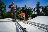 Two men standing on a roof measuring tracks to install solar panels, palm trees behind them