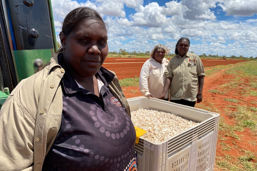 A First Nations woman stands next to a bin of garlic.  There are two women standing behind her, and blue sky is visible