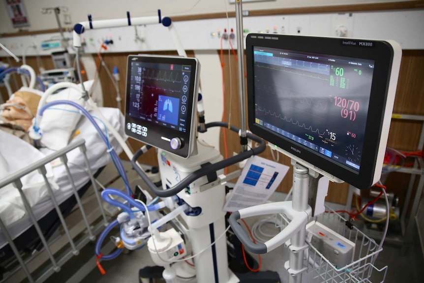 A close-up shot of ventilator monitors in a hospital room with a bed in the background and a dummy's head on a pillow.