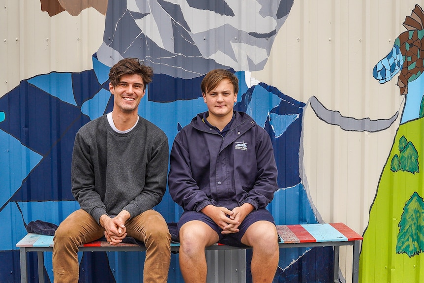 A young male teacher and male student sit on a colourful outdoor bench smiling.