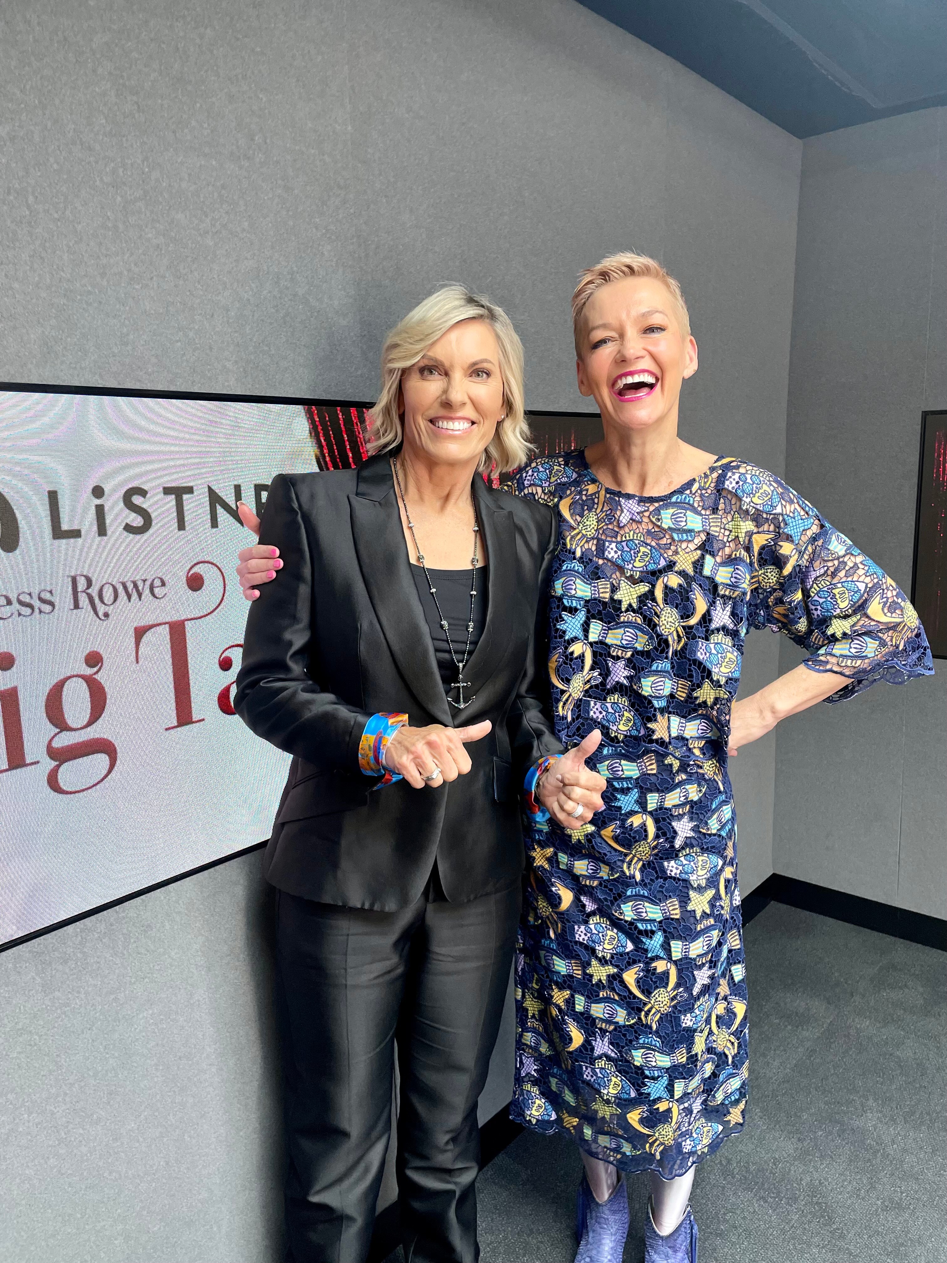 Two blonde white women stand smiling, one's arm around the other. One is in a black suit, the other a colourful dress.