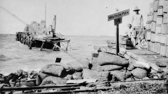 Old photo of man standing on pier with sandbags, sign reads "Watson Pier"