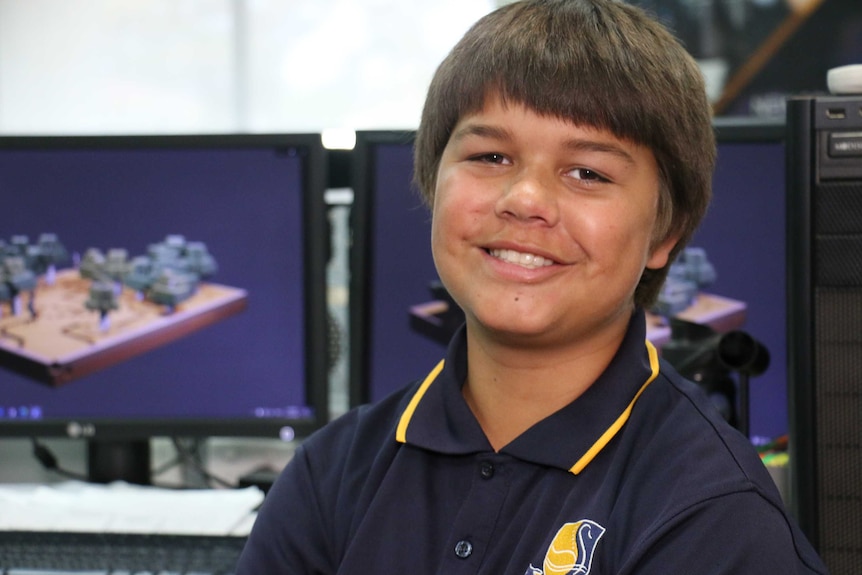 A boy wearing a polo shirt with a school logo on it smiles at the camera in front of two computer screens.