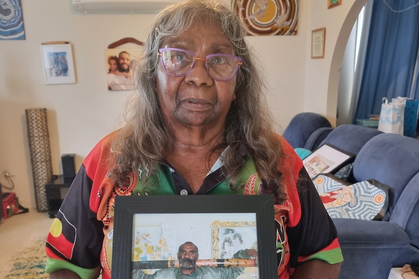A woman stands in a lounge room holding in front of her a photo of a man carrying a painted didgeridoo
