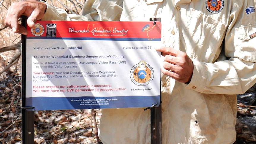 A close-up of a man whith his head unseen holding a red, white and blue outdoor sign for visitors to Wunambal Gaambera country.