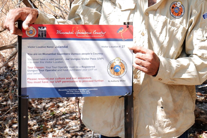 A close-up of a man whith his head unseen holding a red, white and blue outdoor sign for visitors to Wunambal Gaambera country.