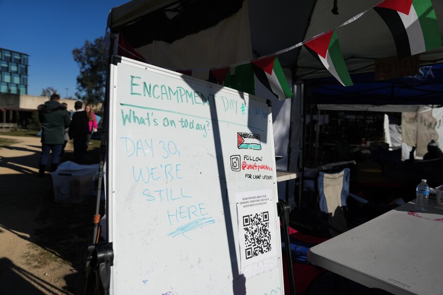 A whiteboard sign saying "Day 30, we're still here" stands next to a tent.