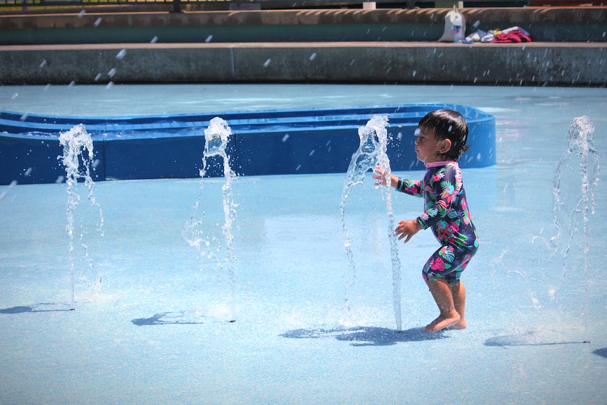 A toddler runs through a water fountain in front of a waterslide