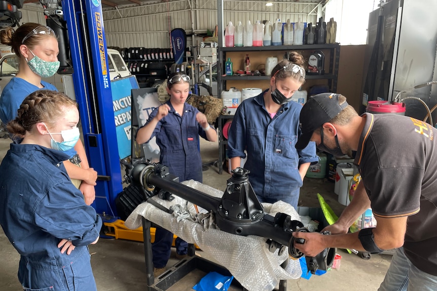 Four girls and a man working on a car axle.