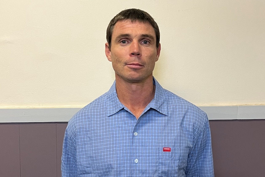 A mean in a blue shirt stands in front of a wall. 