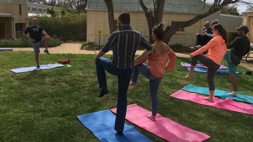 A group of people in workout gear prepare for tree pose during a yoga class. An instructor stands in front of the group