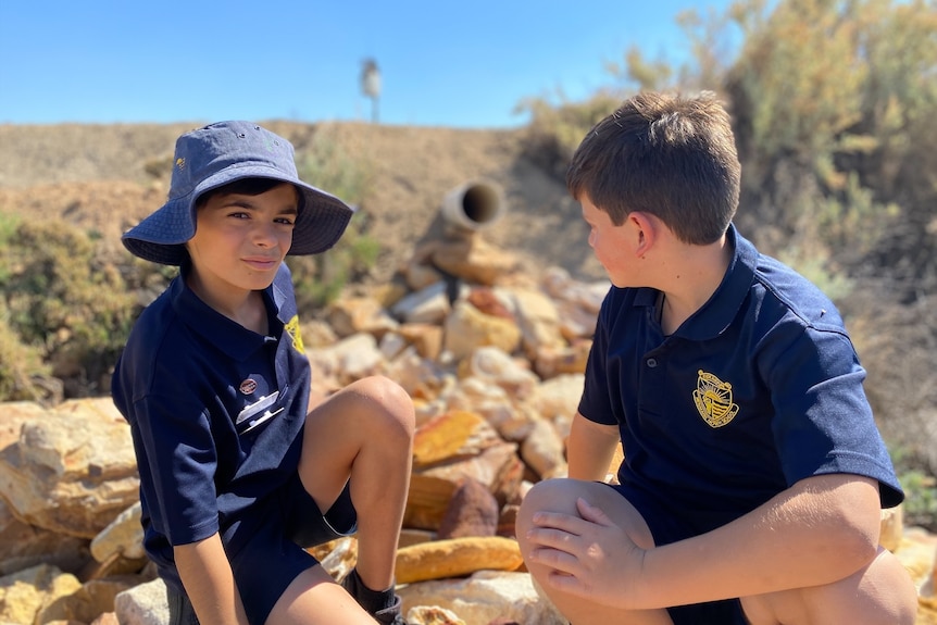 Two boys sit either side of a water pipe on some rocks. One is looking at the camera, one is looking at the pipe behind.