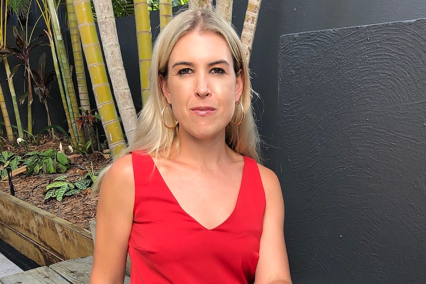 Blonde woman with gold earrings Lexi Crouch sits on a wooden step staring seriously at camera