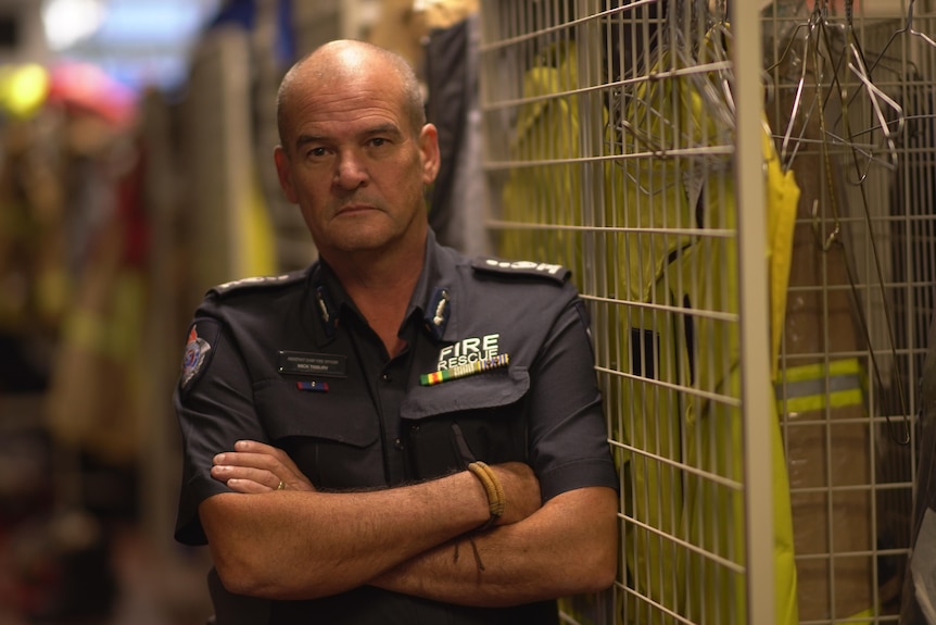 A firefighter in navy formal uniform shirt stands with arms crossed next to uniform cage at a fire station