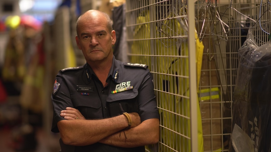 A firefighter in navy formal uniform shirt stands with arms crossed next to uniform cage at a fire station