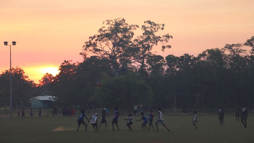 Young boys play football on an oval at sunset.