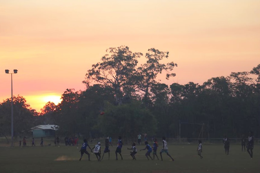 Young boys play football on an oval at sunset.