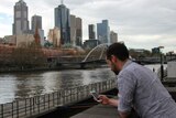 A man leans on a black stone wall and looks at his phone. Melbourne's Yarra River and CBD are in the background.