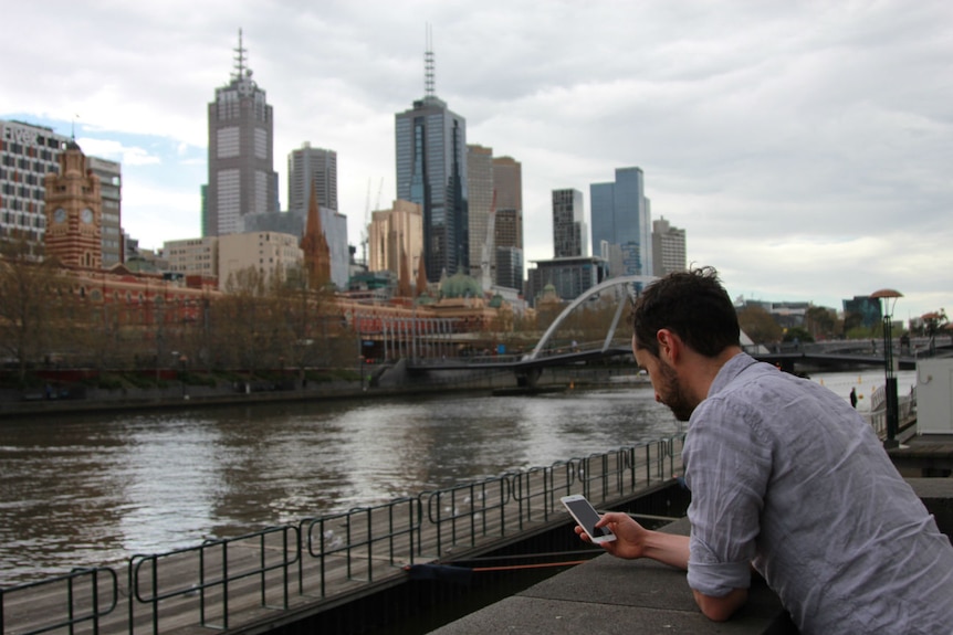 A man leans on a black stone wall and looks at his phone. Melbourne's Yarra River and CBD are in the background.