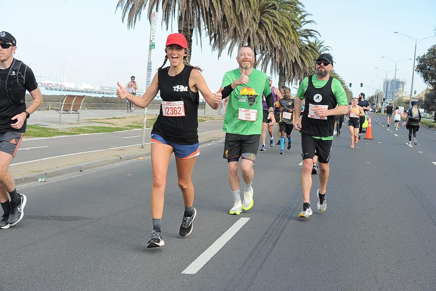 An image of a woman smiling with her thumbs up while running a marathon, with other runners visible in the background. 