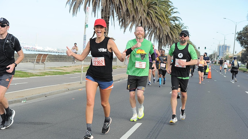 An image of a woman smiling with her thumbs up while running a marathon, with other runners visible in the background. 