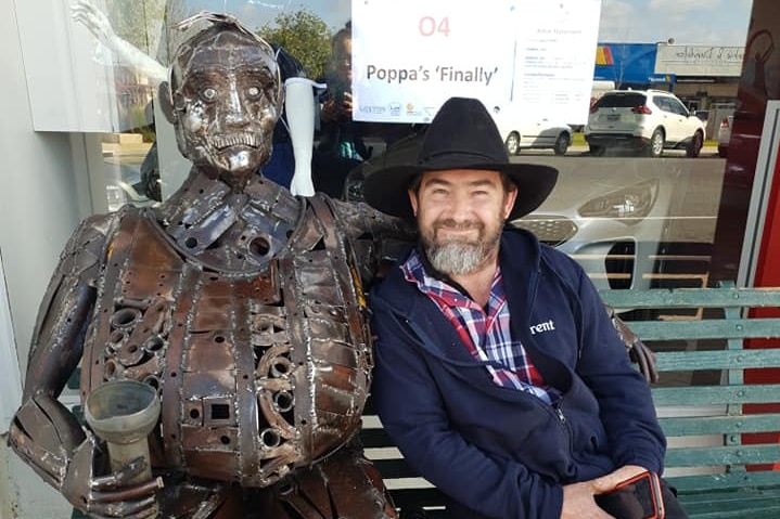 A man in a hat is sitting next to a metal sculpture he has made of a man holding a cup.