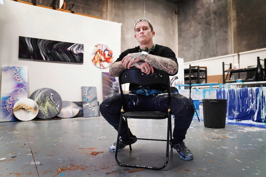 An ex-AFL footballer sits astride chair with a series of his artworks behind him in the background.