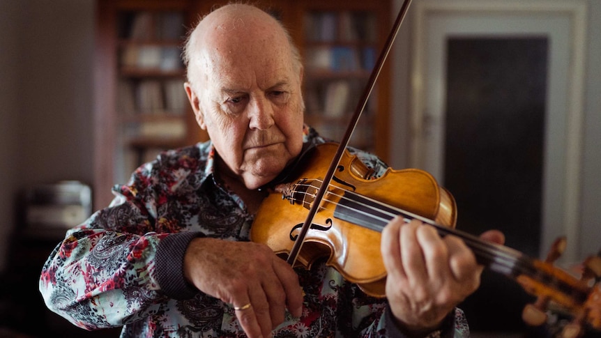 A bald man wearing a dark floral shirt playing a violin