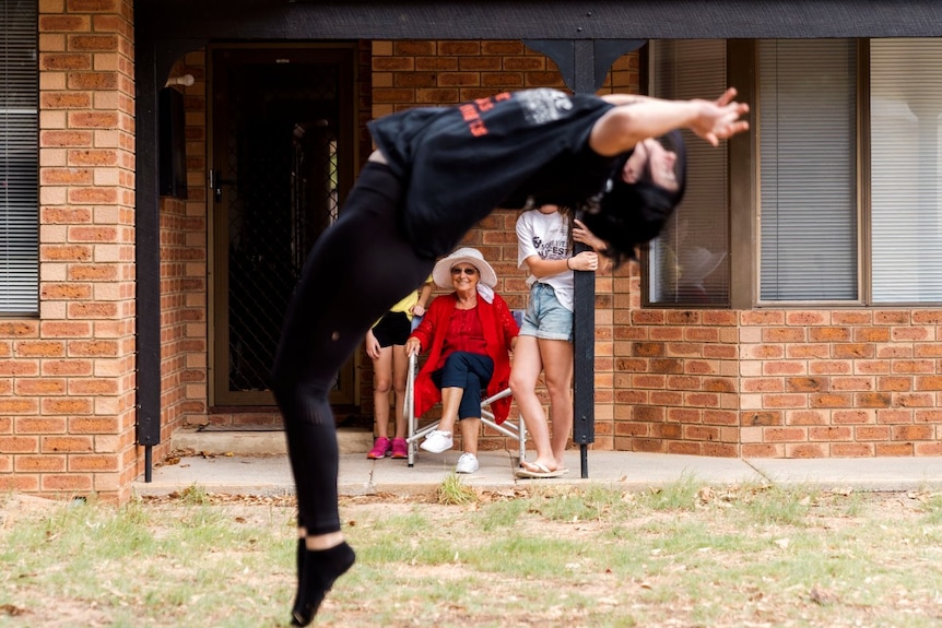 A teenage girl performs a backflip while her grandmother watches on from a chair.