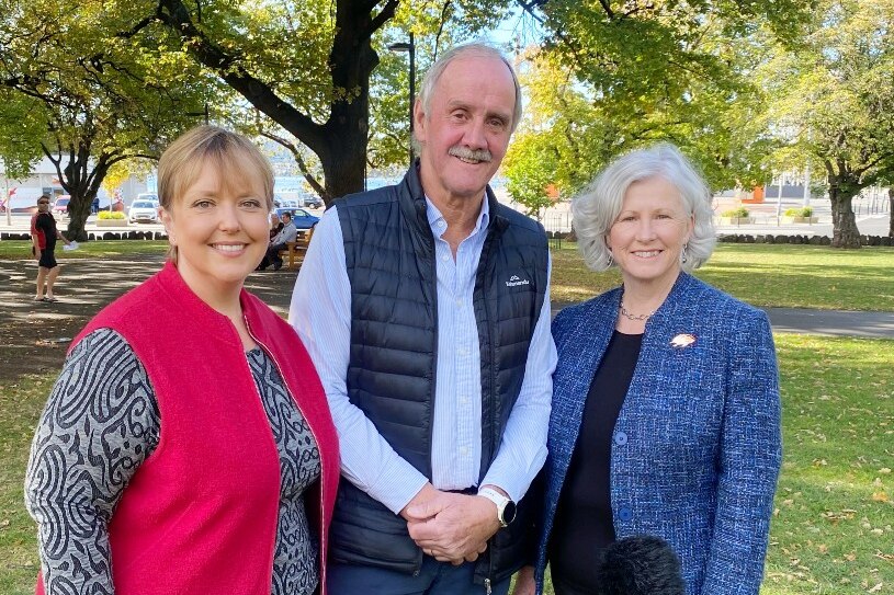 A tall balding man stands in a park flanked by two women