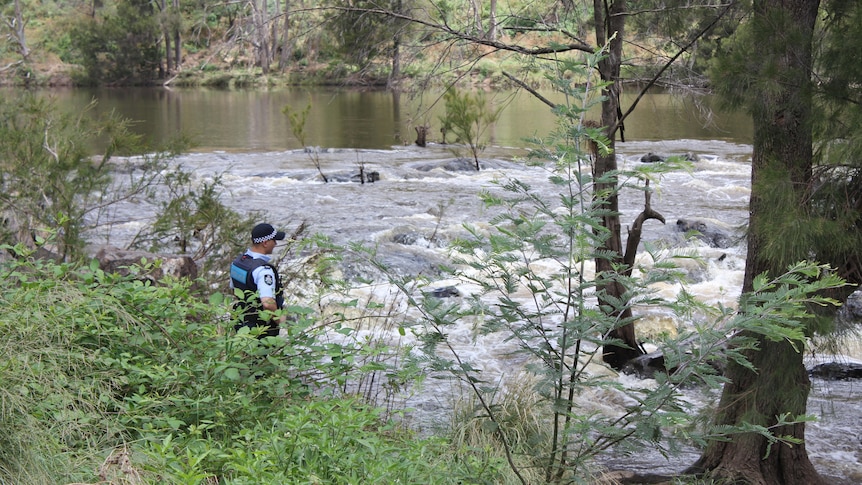 A police officer looks out at the river from the shore, trees all around.