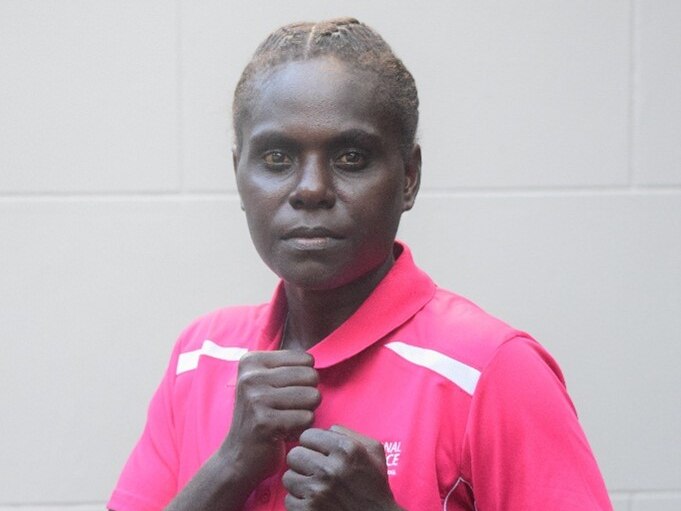 A female boxer from Papua New Guinea is wearing a pink polo shirt, has her fists raised, and is staring at the camera.