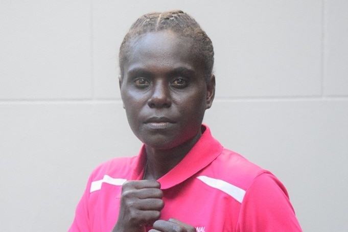 A female boxer from Papua New Guinea is wearing a pink polo shirt, has her fists raised, and is staring at the camera.