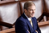 Politician Jim Jordan wearing blue suit stands with arms folded by himself amidst empty brown leather chairs