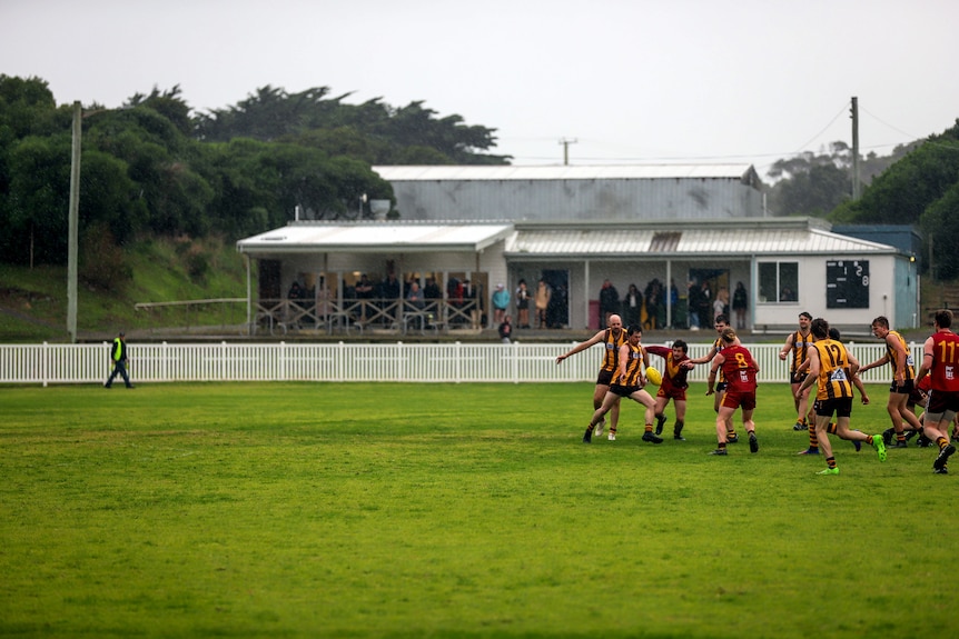 A rainy mid afternoon country football game with two sides in the middle of an oval and a small number of spectators sheltered 