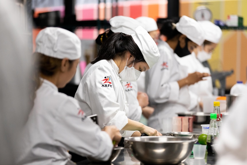 A row of people in chef uniforms cooking and preparing on a stainless steel bench.