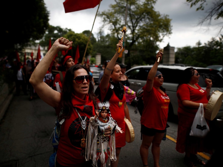 Indigenous rights activists march in Toronto on Canada Day. They are carrying flags, instruments and a doll in native dress.
