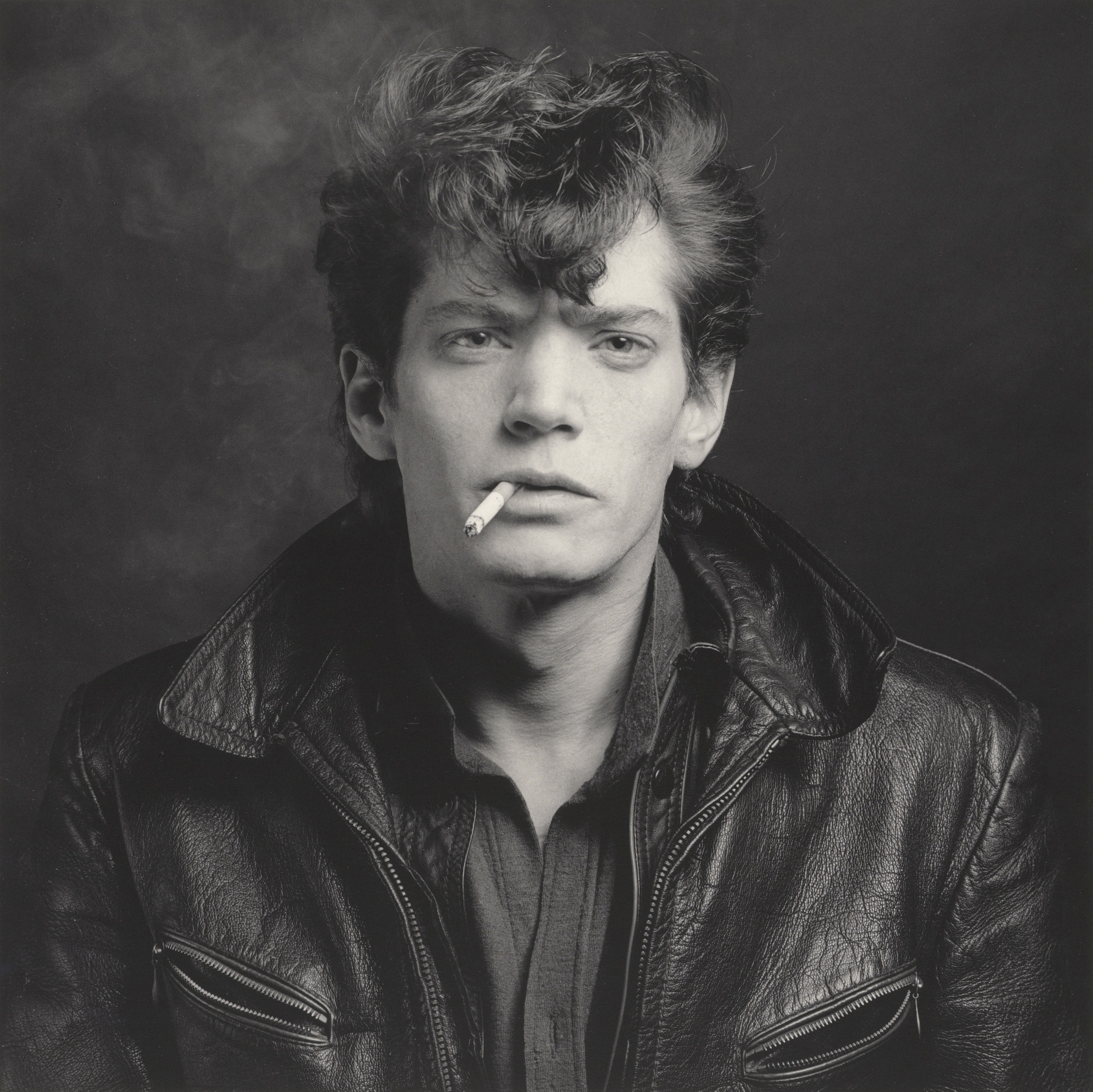 Black and white square format photograph of Robert Mapplethorpe smoking a cigarette in front of a studio background.