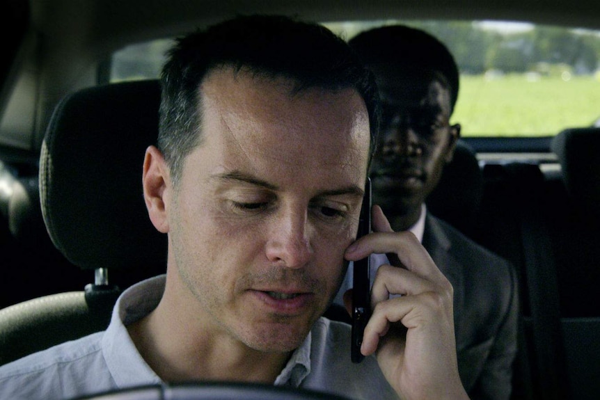 Two men sit in a car, the first in the drivers seat with a phone held to his ear, the second in the back seat looking anxious.